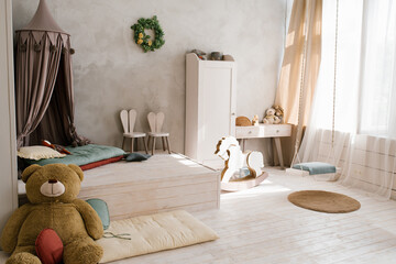 Fototapeta na wymiar A large stuffed bear toy in the interior of a children's room with a teepee. Wooden horse gurney, bunny chairs and wardrobe