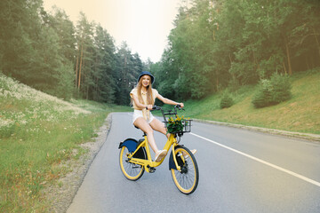 An attractive young tourist rides a bicycle on a road near the forest, stretching her legs and having fun and excited expressions, in the open air.