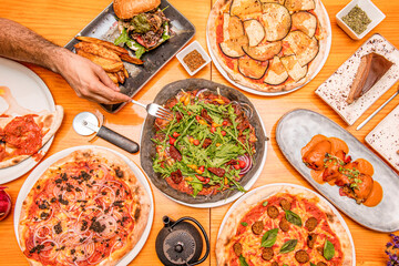 Plates of Spanish food and Italian pizzas viewed from the top with a charcoal flour dough pizza in the center and a man's hand with a fork ready to carve