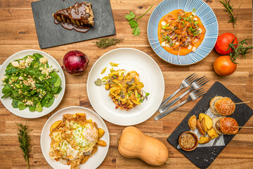 Top view view of delicious Mediterranean food dishes on wooden table. Fried pasta, spinach and pine nut salad, fried eggs with potatoes. Pumpkin cream, green tomatoes