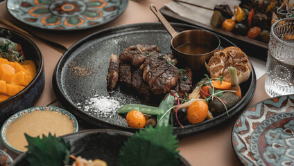 Cooked medium raw beef steak with blood sauce salt and pepper on black rough clay plates served with roasted garlic, peas, tomatoes and carrots. Flat lay between other dished and clutter ready to eat