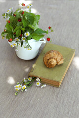 A bunch of strawberries and daisies in a teapot next to an old book and a sink on a table in the garden. Vertical photo background. Tranquil summer vacation and relaxation concept