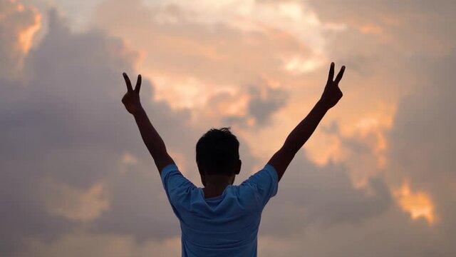 Portrait of an Indian kid making peace sign in front of the orange clouds during the sunset. Kid looking at the sunset clouds and finding peace. Happy kid makes V sign with his hands raised.