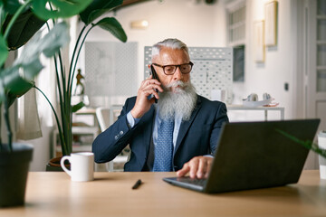 Senior business man doing call with mobile phone while working with laptop computer inside modern office - Focus on face