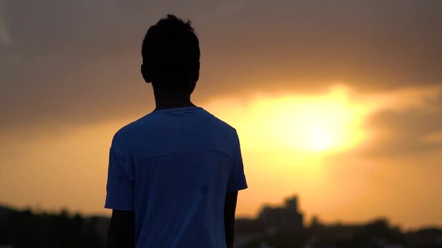 Silhouette of an Indian kid looking at the orange sky during the sunset. Hopeful kid stares at the sun. Kid lost in deep thoughts after looking at the sunset. 
