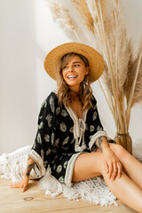 Laughing blond woman in straw hat and boho dress  posing in studio over white background  with pampas grass decor.  Pillows with macrame ornament. Summer trends. © Svetlana Sokolova