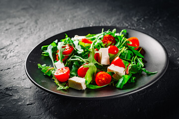 Fresh salad with tomatoes, arugula and cheese cubes on black plate on Dark grey black slate background