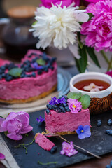 Obraz na płótnie Canvas A piece of berry cheesecake with a broken piece of a spoon lying next to a mug of tea, violet flowers and on a background of peonies and a whole cake