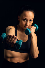 Vertical portrait, in studio, in front of a black background, of an Asian fighting athlete, Korean, boxing with weights in her hand, looking at the camera, with a serious, determined expression.