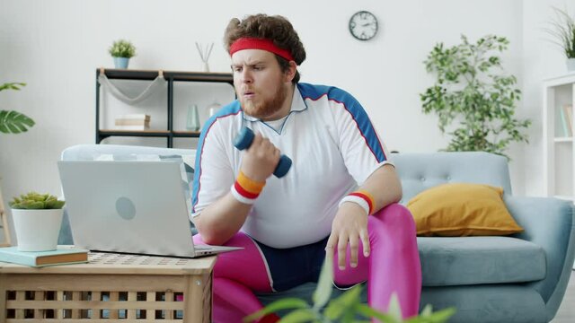 Overweight guy is watching sports tutorial with laptop exercising with dumbbell then eating pizza sitting on couch at home. Technology and people concept.