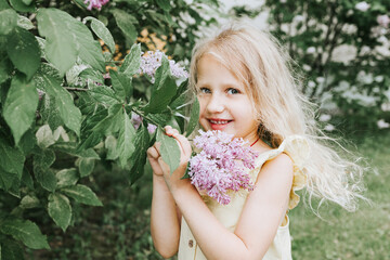Naklejka na ściany i meble portrait of a thoughtful and sad little blonde girl in a yellow dress standing, smelling flowers near a blooming lilac in spring, the concept of a happy childhood and school holidays