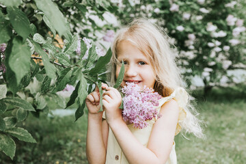Obraz na płótnie Canvas portrait of a thoughtful and sad little blonde girl in a yellow dress standing, smelling flowers near a blooming lilac in spring, the concept of a happy childhood and school holidays