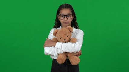 Portrait of cute unhappy African American teen girl hugging teddy bear standing on green screen. Sad nerd teenager in eyeglasses posing at chromakey background looking at camera - Powered by Adobe