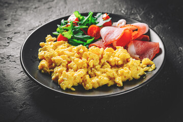 scrambled eggs with prosciutto and salad on black plate on Dark grey black slate background