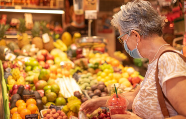 Senior woman wearing surgical mask visit a traditional food market choosing some fruit, holding a glass of watermelon in pieces