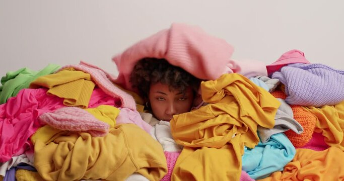 Unhappy tired disorganised woman covered with big heap of multicolored clothes drowned in laundry cluttered with clothing poses against white background. Closet cleaning concept. Disorder and mess