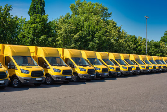 A Lot Of Sustainable Electric Yellow Delivery Vans Are Parking In Front Of Green Trees.