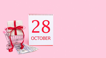 A gift box in a shopping trolley, dollars and a calendar with the date of 28 october on a pink background.