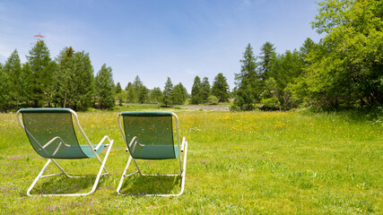 Two beach chairs from behind for leisure in a natural peaceful environment, with grass and forest and blue sky in the background. With copy space