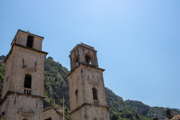 Fototapeta na wymiar Church towers visible against the backdrop of a mountain landscape and green trees. Old medieval church in backdrop of the mountains in Kotor Montenegro