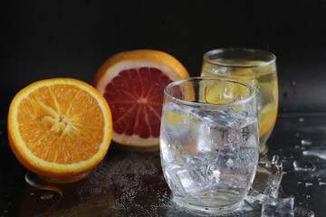 Ice cube flies falls into the takan with water soda next to lie orange grapefruit and ice on a black background. Refreshing drinks. Lemonade, fruit water