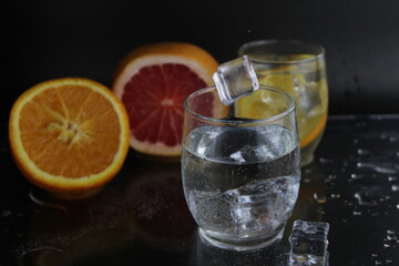 Ice cube flies falls into the takan with water soda next to lie orange grapefruit and ice on a black background. Refreshing drinks. Lemonade, fruit water
