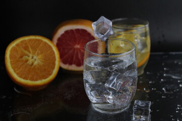 Ice cube flies falls into the takan with water soda next to lie orange grapefruit and ice on a black background. Refreshing drinks. Lemonade, fruit water