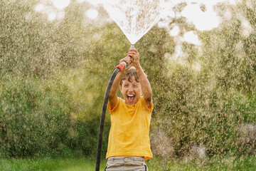happy boy splashes and plays with garden hose with sprinkler at sunset in backyard, enjoys...