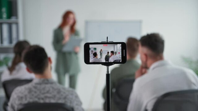 remote meeting, young woman conducts training to business team showing presentation on flipchart during video broadcast using mobile phone in office