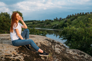 Nature, wellbeing, Relaxation, meditation and mental health. Interacting, Thriving with nature. Calming and enchanting effect. Young redhead woman sitting on rock and watching on river on sunset