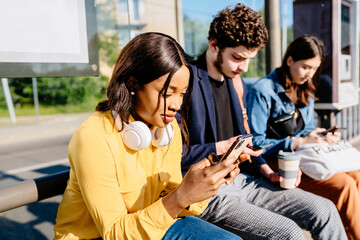 Group of three multiethnic friends using smart phone while sitting on the bench at bus stop and waiting for public transport at sunny bright summer day.