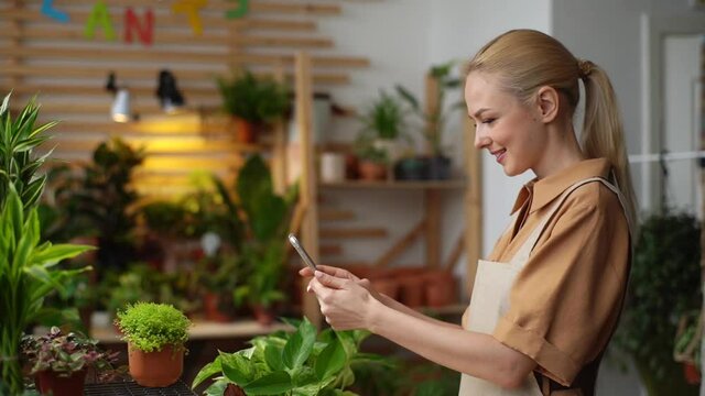 Side view of cheerful female florist and blogger taking pictures of plants in floral shop using mobile phone camera. Happy female blogger or vlogger is review small tree in pot on social media.
