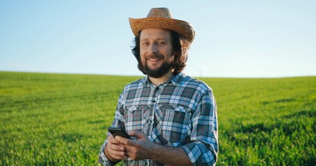 Naklejka na ściany i meble Waist up portrait view of the optimistic caucasian farmer scientist in hat using smartphone mobile technology application while working at the field and smiling. Agriculture concept