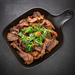 Thai food, fried sun dried beef with crispy basil leaves in black pan on the dark tone texture background
