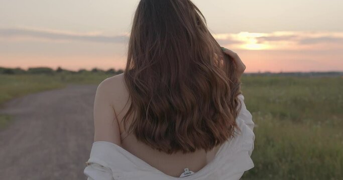 Back view of romantic woman in summer dress walking at countryside and enjoying amazing sunset. Leisure time on fresh air. Natural beauty.