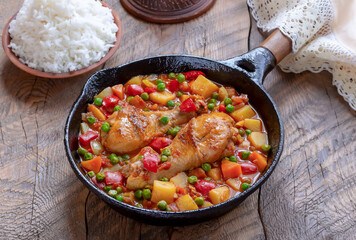 Chicken Afritada. Braised Chicken in Tomato Sauce mixed with potatoes, carrots, bell pepper, and green peas served in frying pan with bowl of with cooked rice on background. 
