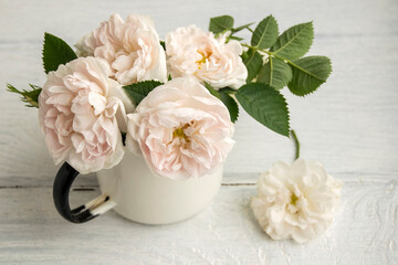Delicate pink roses are in the old cup. The cup is on a white wooden table.