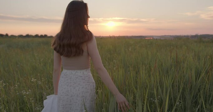 Back view of young woman with brown hair wearing romantic dress walking on field during summer sunset. Concept do people and freedom.