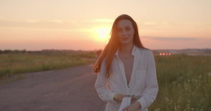 Pleasant Young Woman With Wavy Brown Hair Wearing Stylish Summer Clothes Walking On Field. Playful And Romantic Mood. Amazing Sunset On Background. 