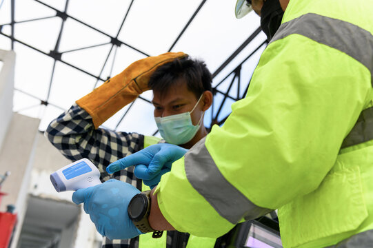 Labour Screening, Construction foreman wearing protective face mask pointing measurement results of body temperature of welder worker in an Infrared Thermometer during coronavirus COVID-19 Pandemic