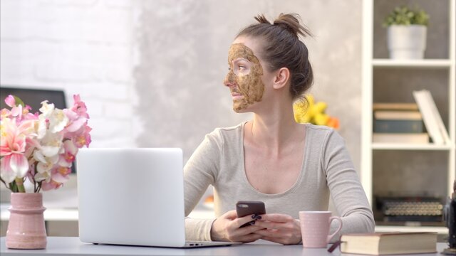 Woman Cosmetic Facial Skin Care. Woman In Skin Rubber Peeling Face Beauty Mask Sitting At Desk Using Phone And Laptop.