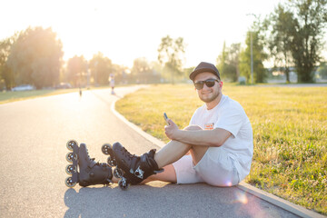 Young smiling man, relaxing, wearing inline skates. Taking a selfie, with his phone, sitting on ground.