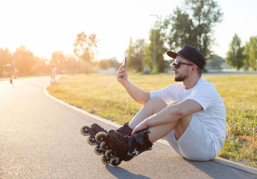 Young smiling man, relaxing, wearing inline skates. Taking a selfie, with his phone, sitting on ground.