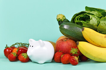 Concept for costs of healthy eating or saving money when buying food. Vegetables and fruits next to piggy bank on blue background with copy space - obrazy, fototapety, plakaty