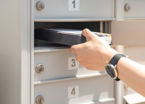 Mail Man Delivering Small Parcel In Mailbox By House Number, Selective Focus. Person’s Hand Holding Package Put In Metal Storage. People Get Packet From Opened Cabinet Door. Sender, Receiver Concept.