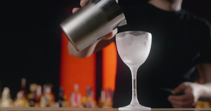 Bartender pours mixed drink from the shaker to the frozed high-stemmed glass, barman makes cocktail with ice and mixed alcohol, 4k 120 fps Prores HQ