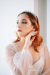 portrait of a charming bride with red hair in a white wedding dress with pearl accessories on her...