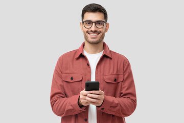 Young modern man holding smart phone in hands, looking at camera with smile, isolated on gray...