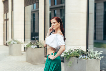 Portrait of a red-haired girl in a summer blouse and skirt against the background of a modern building