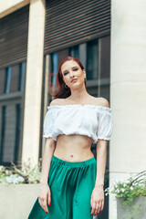 Portrait of a red-haired girl in summer posing at the camera against the background of a modern building